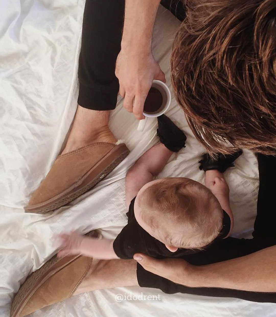 Man sitting on bed with baby wearing EMU sheepskin slippers and holding coffee