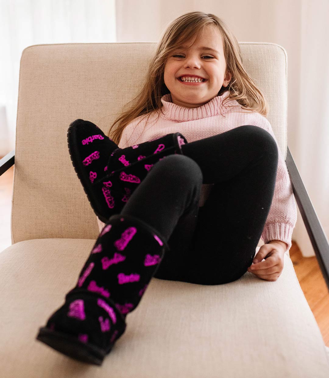 Young girl sitting on seat wearing black sheepskin boots with Barbie logo print and smiling
