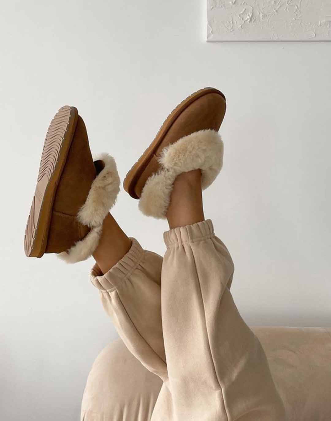 Woman laying on couch with legs up in the air, wearing Chestnut coloured sheepskin slippers and tonal tan trackpants