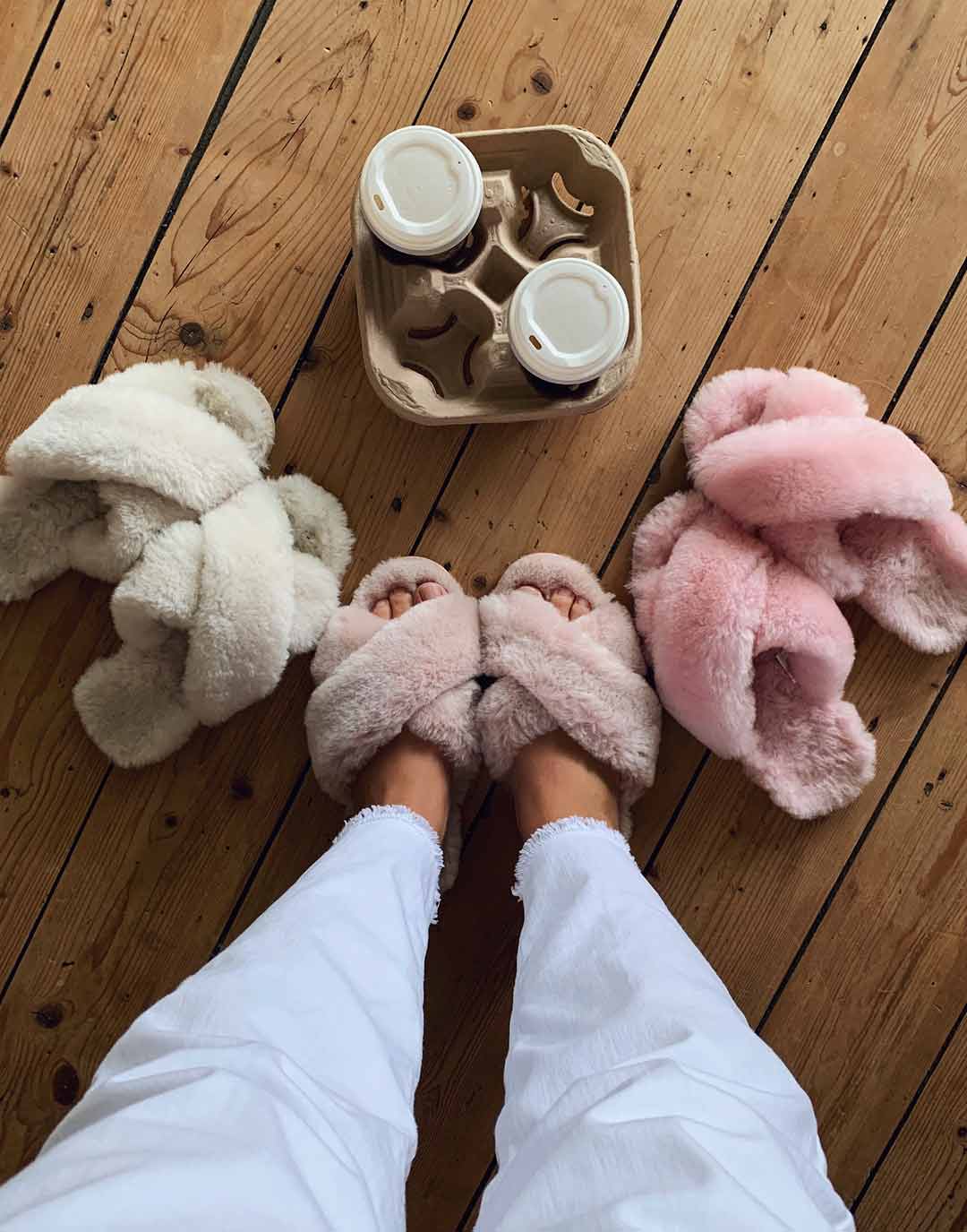 Woman standing on hardwood floors wearing a pair of Musk Pink coloured slippers, with two pairs on either side in Natural and Baby Pink colourways