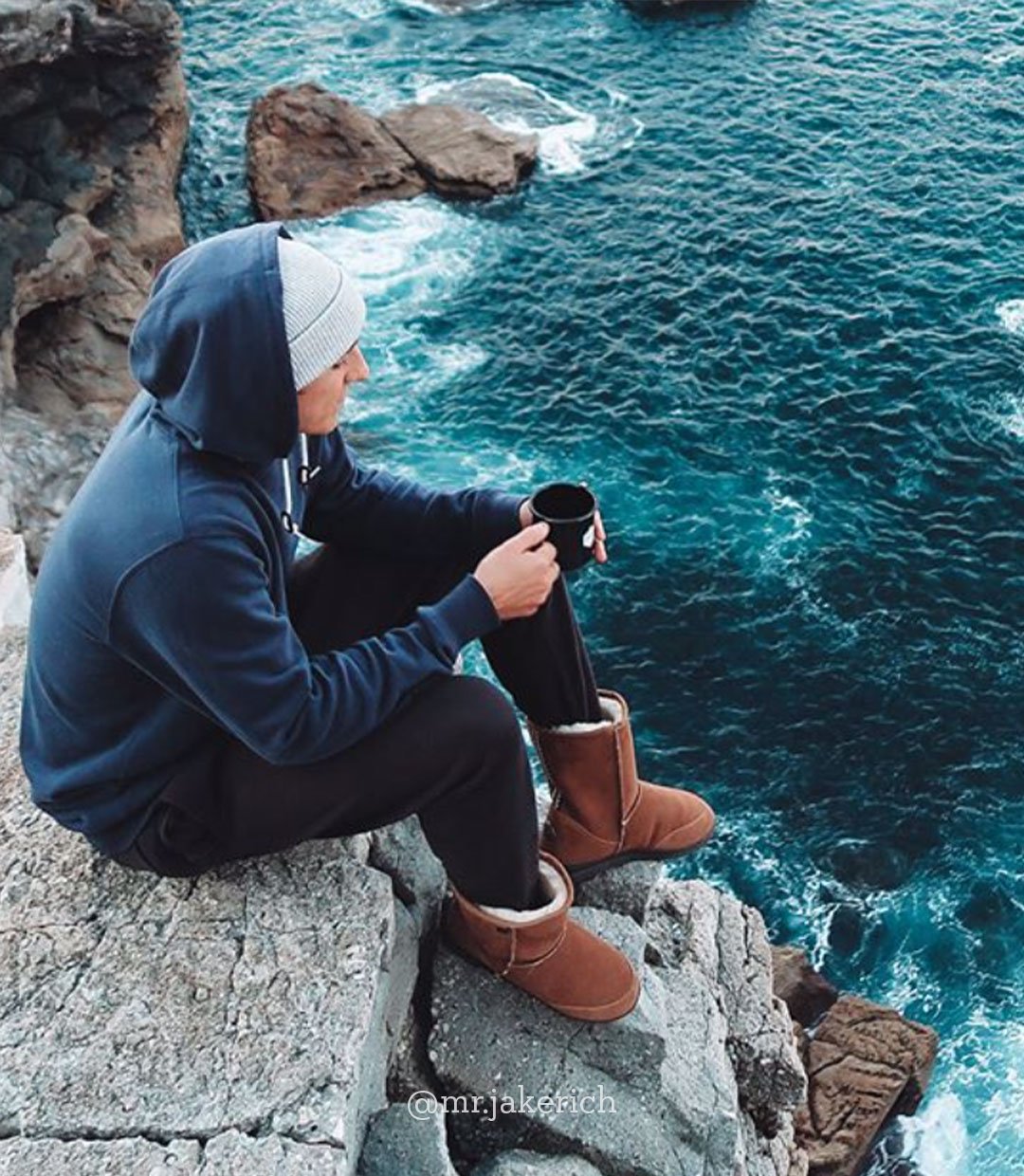 Man sitting on cliff rocks next to ocean holding coffee mug and wearing EMU sheepskin boots