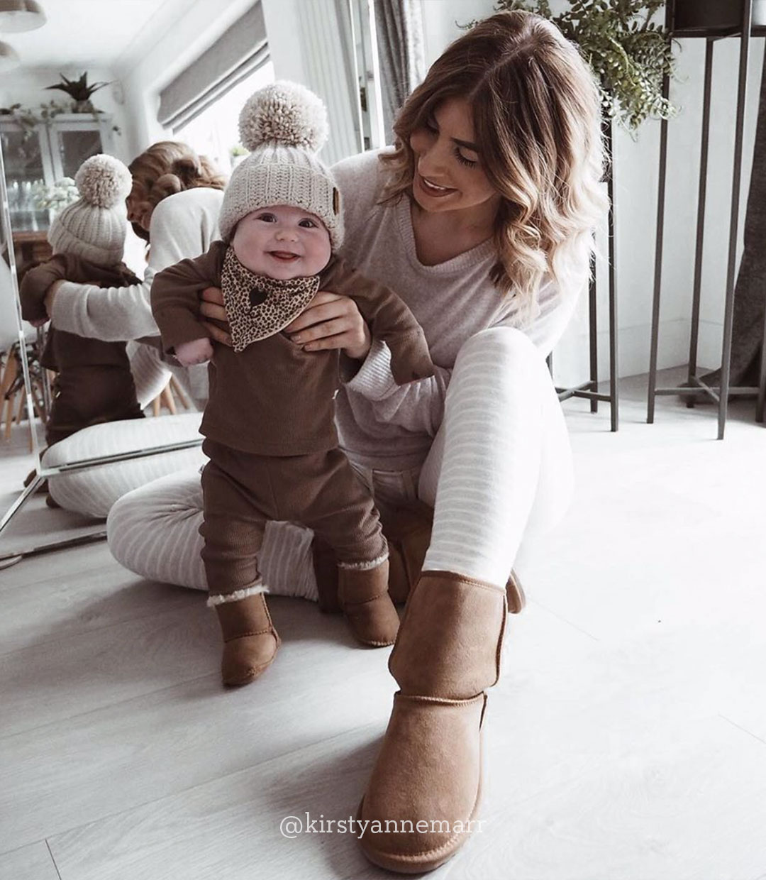 Woman wearing EMU sheepskin boots and sitting on floor holding smiling baby wearing EMU baby booties