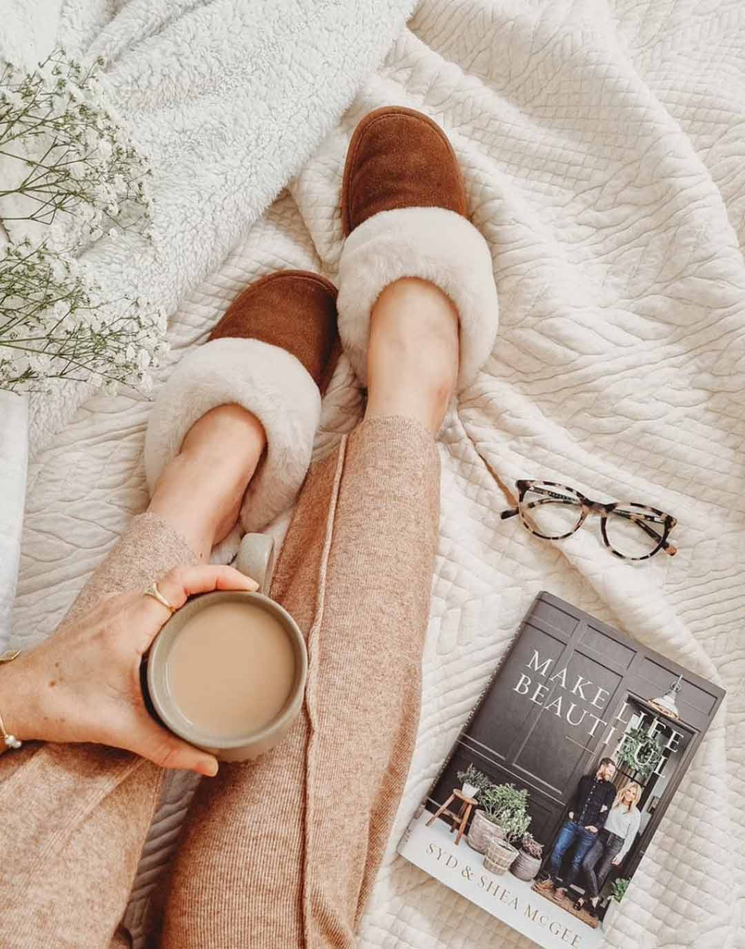 Woman sitting on bed holding a coffee with book and reading glasses next to her, and white quilted blanket wearing mule-style slippers in Chestnut colourway.